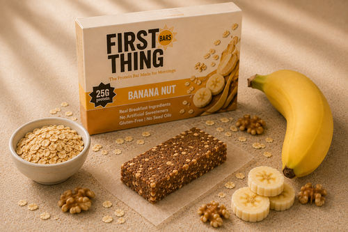 A Banana Nut retail box standing behind one unwrapped bar on parchment, with a bowl of oats, a whole banana, banana slices and walnuts on a tan cloth