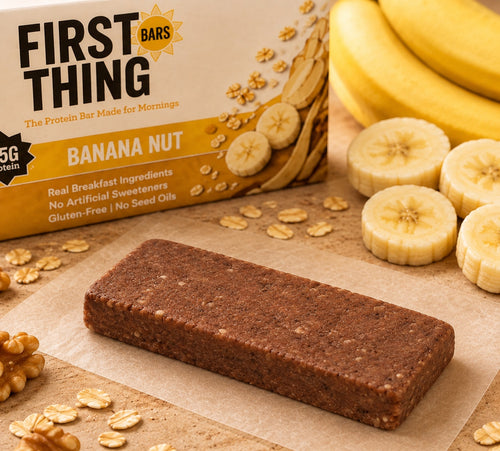One unwrapped Banana Nut bar on parchment in front of a Banana Nut box, with sliced and whole bananas and walnuts