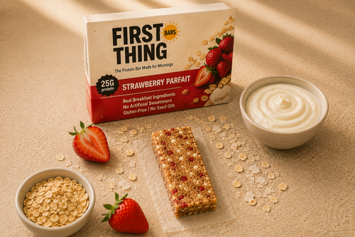A Strawberry Parfait retail box standing behind one unwrapped bar on parchment, with a bowl of yogurt, a bowl of oats and fresh strawberries on a tan cloth