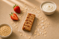 An unwrapped Strawberry Parfait bar on parchment, with fresh strawberries, a bowl of oats and a bowl of yogurt on a tan surface