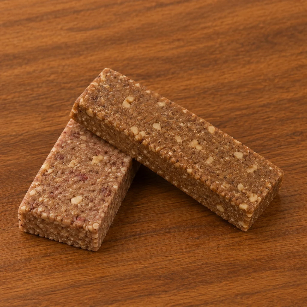 Two First Thing bars crossed on a dark wood table, a pink-flecked Strawberry Parfait bar resting against a Banana Nut bar, close enough to see the nut pieces and seeds pressed through them