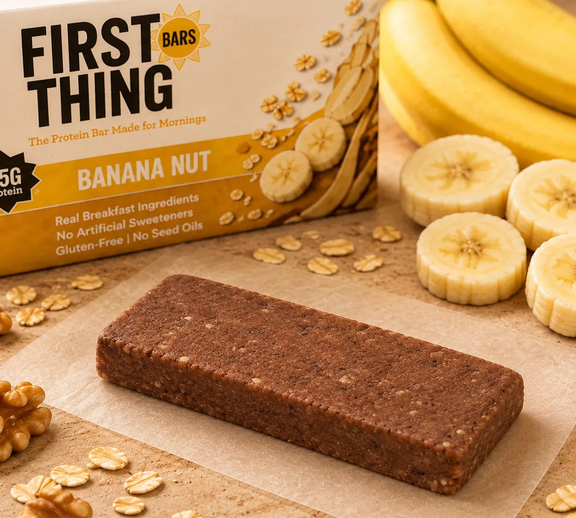 An unwrapped Banana Nut bar on parchment in front of its gold and cream First Thing box, beside a bunch of bananas, banana slices and walnuts