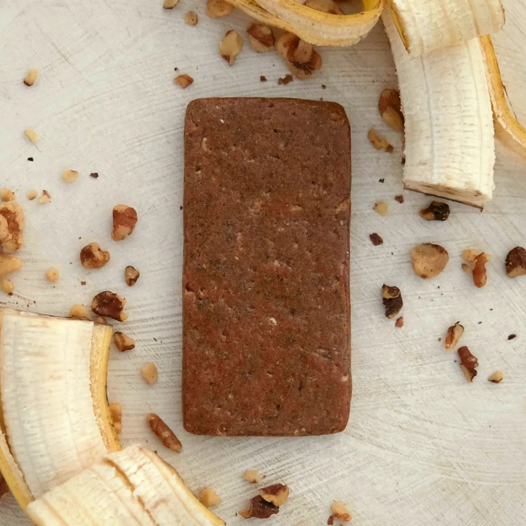 A Banana Nut bar seen from above on a pale wood table, among peeled banana and scattered walnut pieces